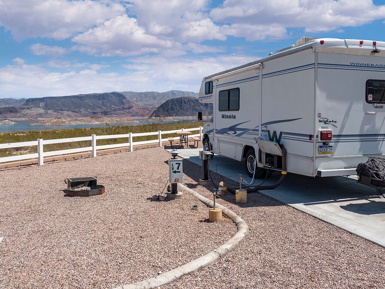 White Winnebago Minnie Class C motorhome parked at a gravel full-hookup site with its power cord and hoses connected, Lake Mead in the background