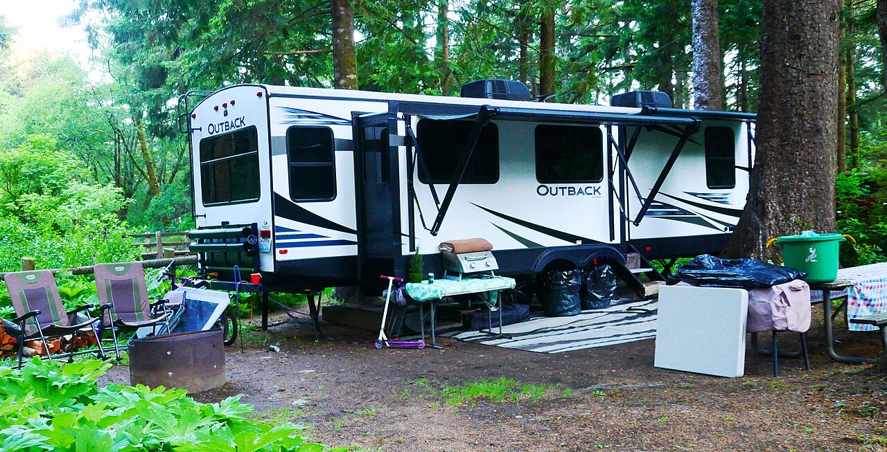 Black and white Keystone Outback travel trailer with its awning out on a forested campsite, camp chairs and a cooler in front