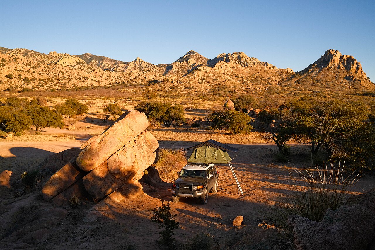 SUV with a rooftop tent parked beside a large boulder at a dispersed campsite on a national forest road, with granite peaks lit by evening sun