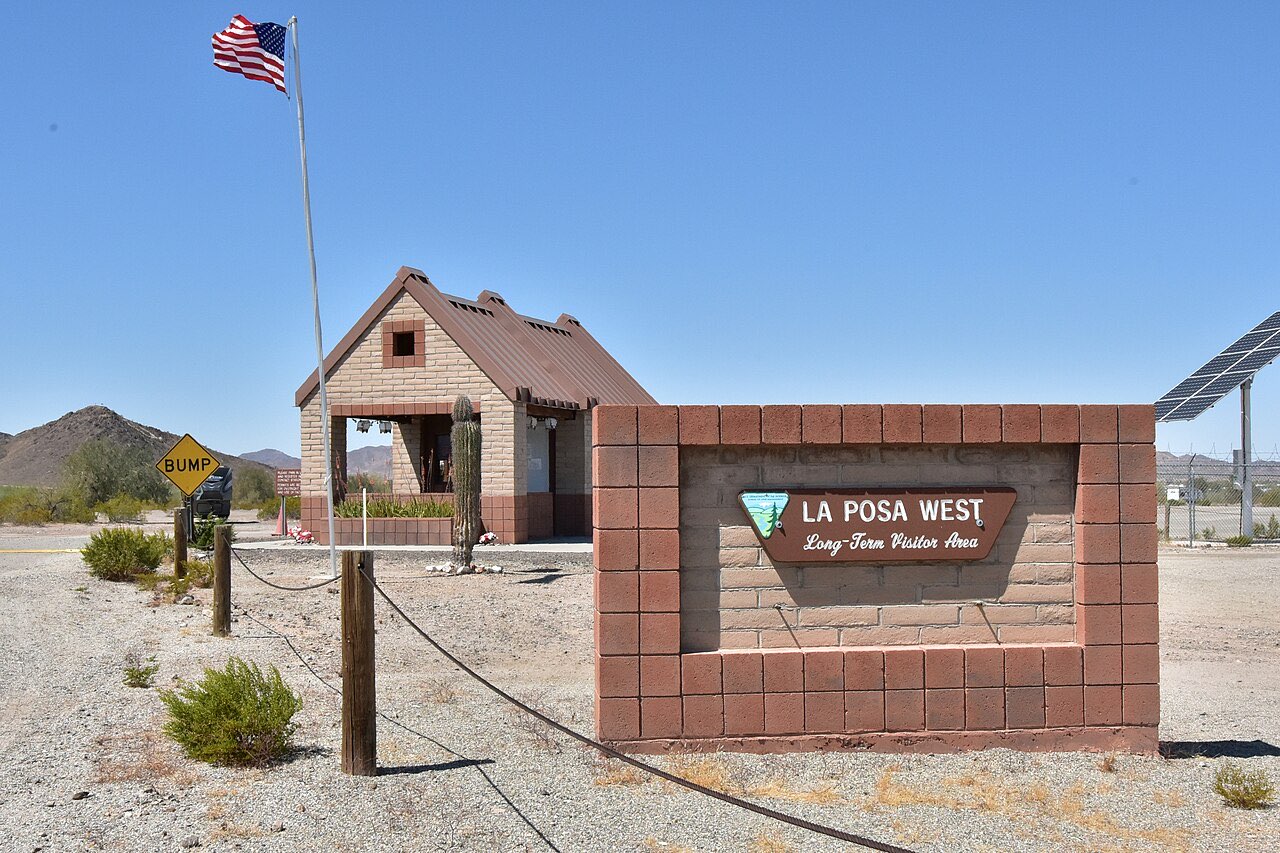 Brown BLM sign reading Please Park in Lot and Register in the Contact Station, Permits Are Required for Overnight Stays, with RVs and desert mountains behind