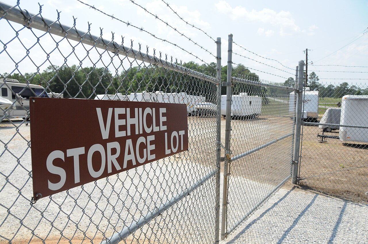 Chain-link gate with a brown Vehicle Storage Lot sign, with travel trailers and campers parked on gravel behind the fence
