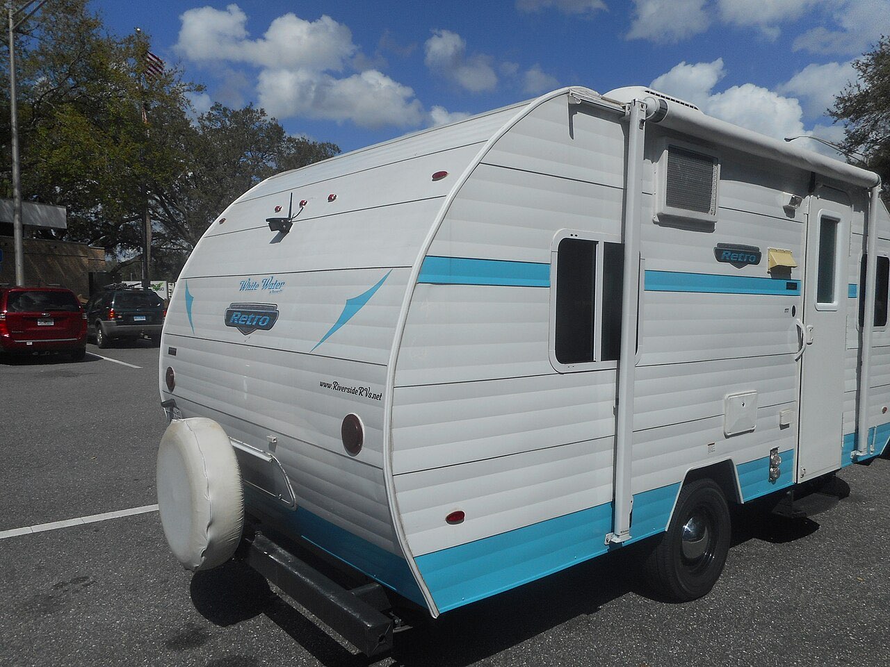 Small white and teal Retro travel trailer with a spare tire on the back, parked at a highway rest area under a partly cloudy sky