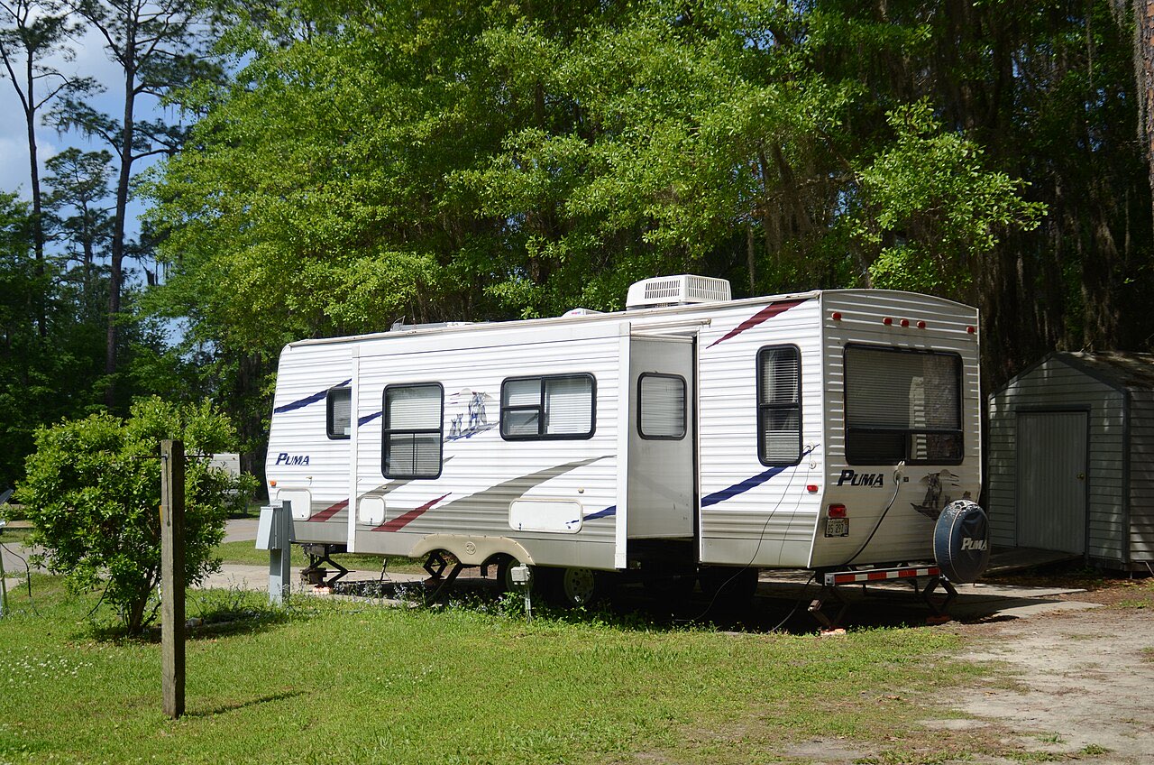 A white Puma travel trailer with a slide-out parked on a grassy campsite under tall pines