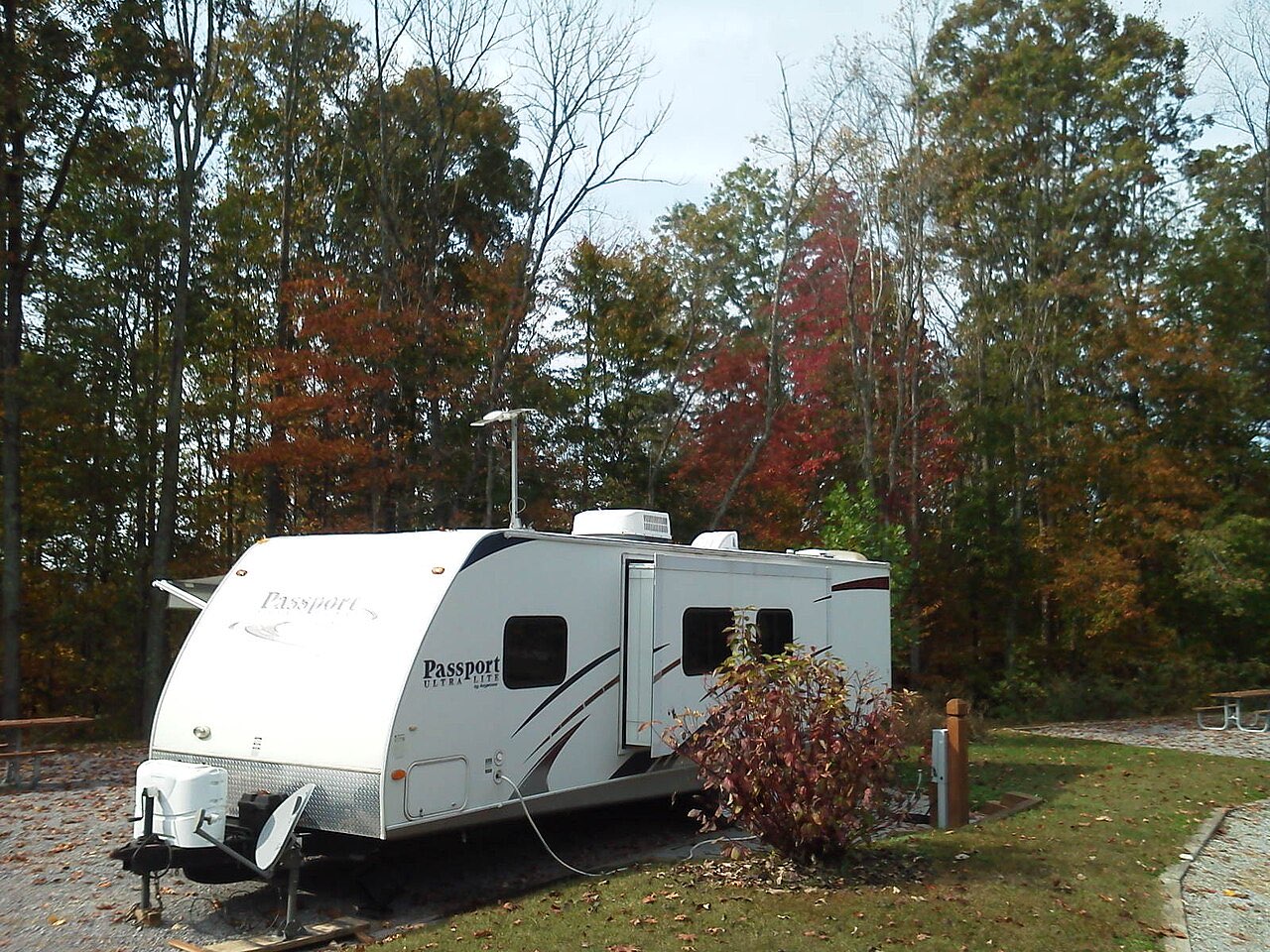 A white Passport travel trailer with a slide-out on a gravel campsite surrounded by autumn trees