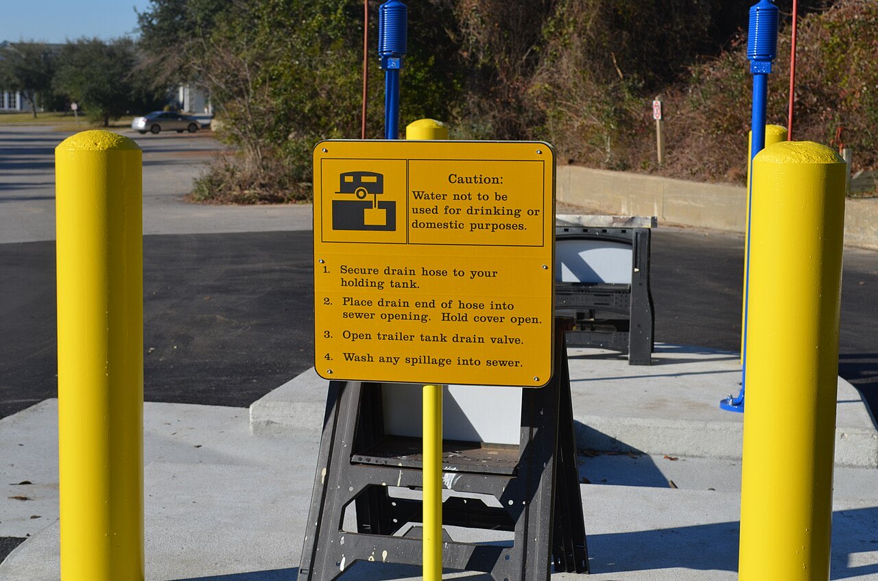 Yellow caution sign at a Virginia state park dump station listing four steps for emptying a holding tank, mounted between yellow bollards over the drain