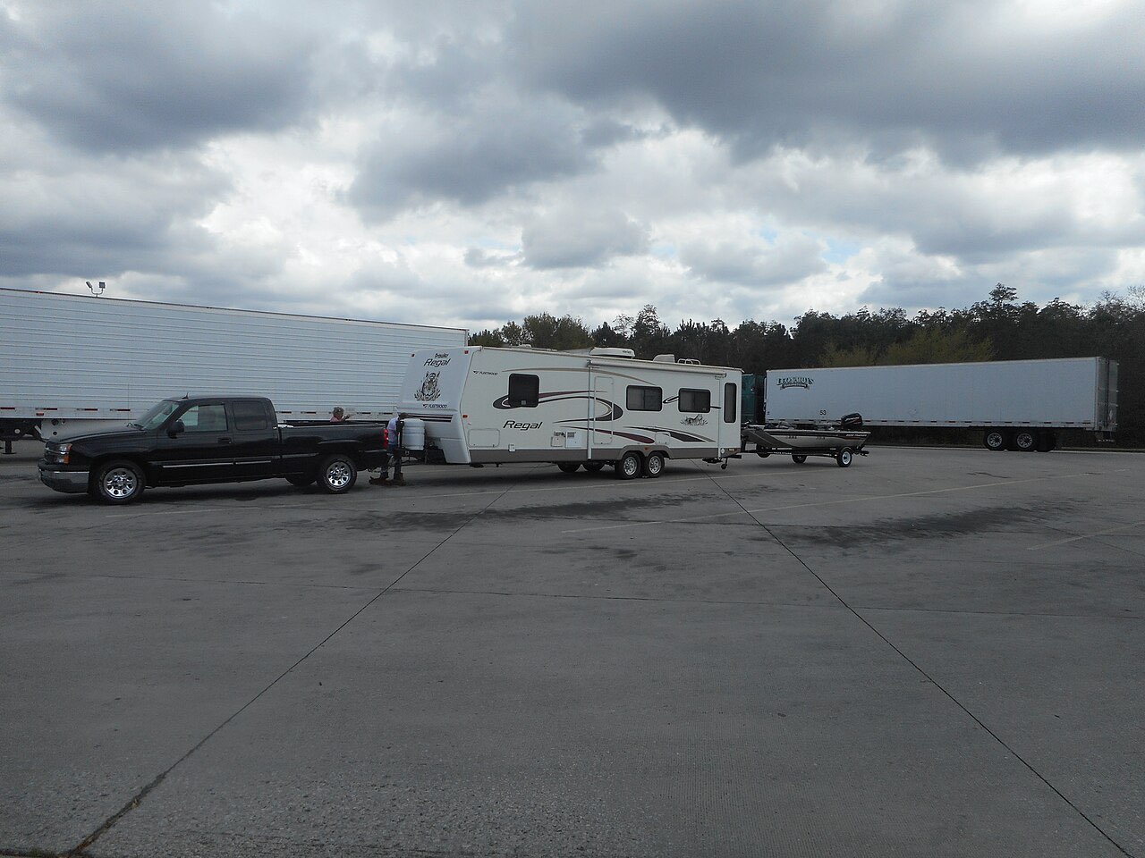 A black Chevrolet pickup towing a white travel trailer with a boat trailer hitched behind it at a highway rest area
