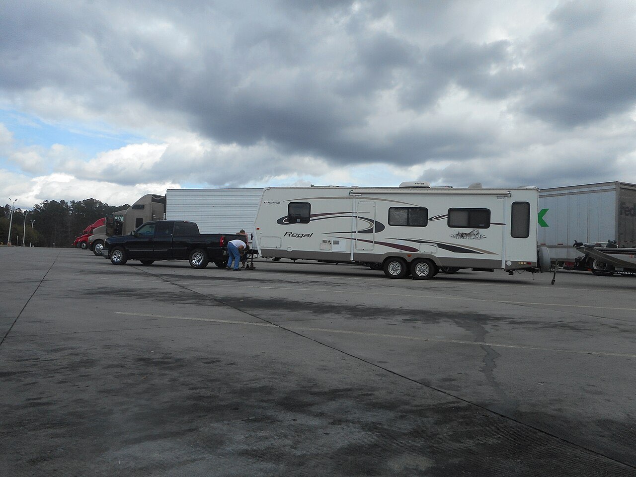 A pickup towing a travel trailer and a boat trailer parked in a rest-area lot beside semi trucks