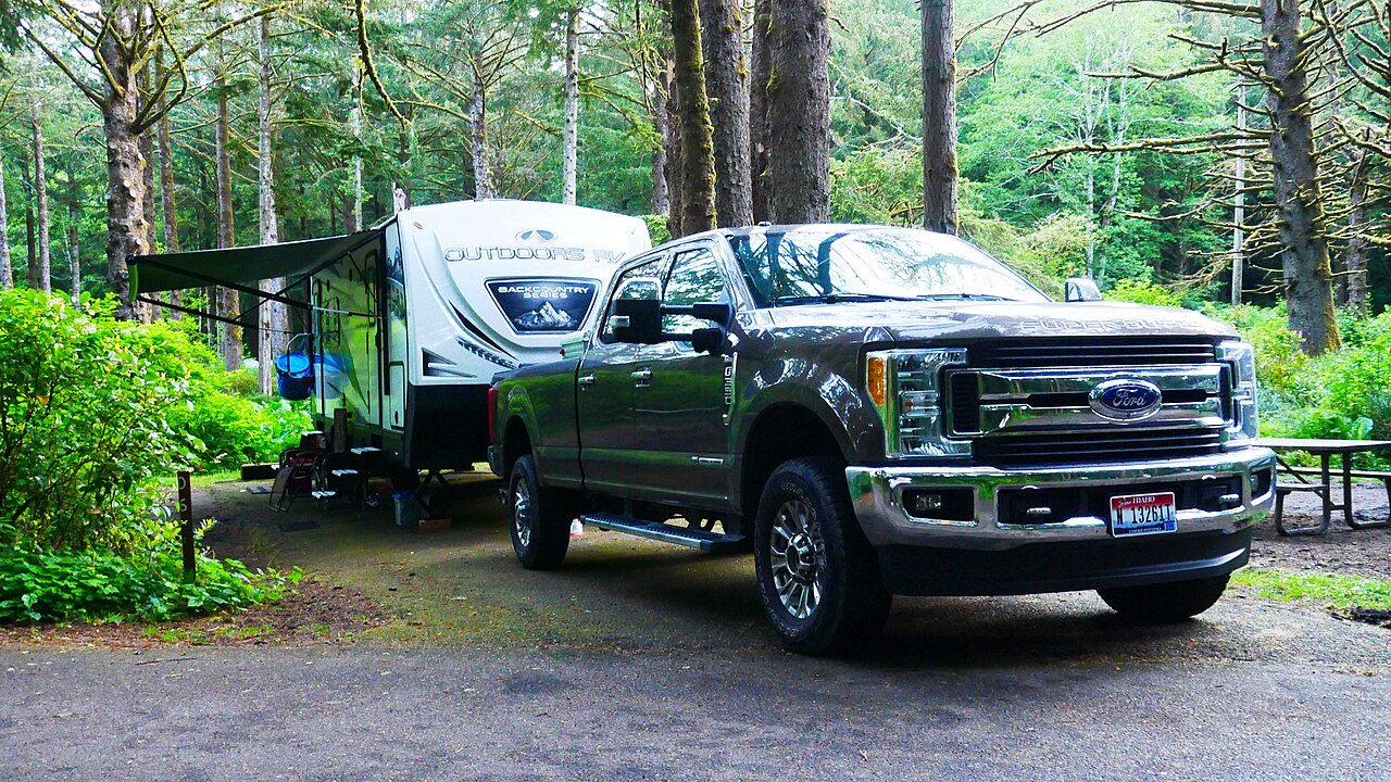 Dark grey Ford F-250 pickup hitched to an Outdoors RV travel trailer on a paved campsite among tall conifers at Beverly Beach State Park, Oregon