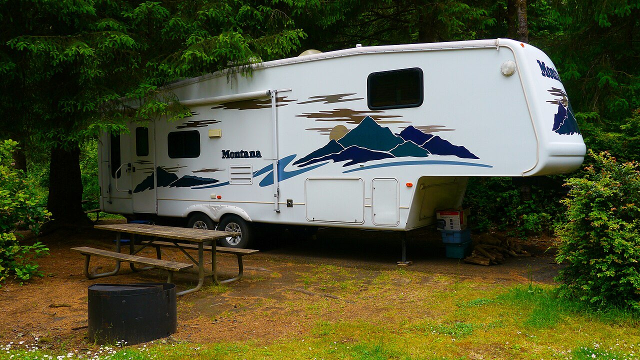 White Keystone Montana fifth wheel with blue mountain graphics parked on a wooded campsite next to a picnic table and fire ring